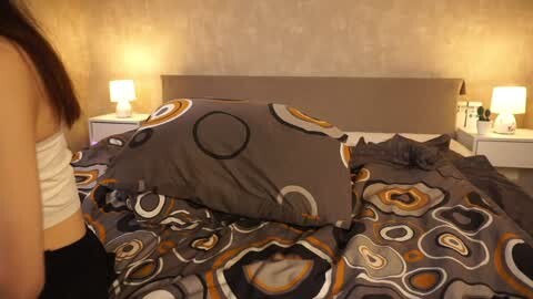 Media: Video of a woman in a white tank top lying on a bed with a gray and brown abstract-patterned comforter, beige wallpaper, and bedside lamps.