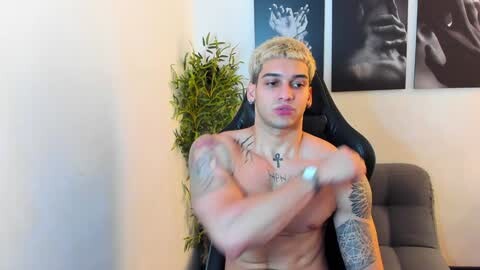 Media: Video of a shirtless, muscular, young man with light skin and bleached blonde hair, wearing a black cross necklace. He's seated in a black gaming chair, surrounded by a potted plant and black-and-white framed art on a cream wall.