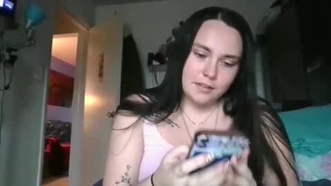 Media: Video of a young woman with long black hair, fair skin, and a tattoo on her left arm, holding a blue phone, in a dimly lit bedroom with a bed, door, and various items visible in the background.