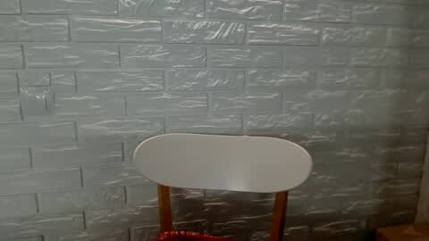 Media: Video of a white, modern chair with wooden legs against a textured, light gray, brick-like wall. The chair's back is smooth and rounded, with a subtle sheen.