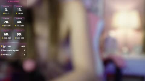Media: The image is a blurred video showing a tall, naked woman with long blonde hair. The background is indistinct, possibly a bedroom with soft lighting. Social media usernames and numerical stats are overlaid on the image.
