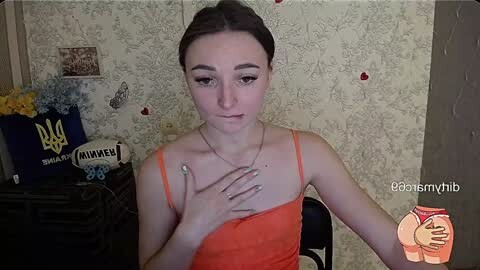 Media: Video of a young woman with fair skin, brown hair tied back, wearing an orange spaghetti-strap top, sitting in a room with floral wallpaper. She has a delicate necklace and is looking at the camera with a slight smile.