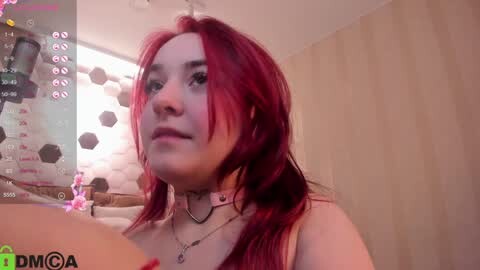 Media: A video of a young woman with long, vibrant red hair, fair skin, and a neutral expression, wearing a white tank top and a black choker. Background shows a cozy, dimly-lit room with a wooden wall and a pink floral pillow.