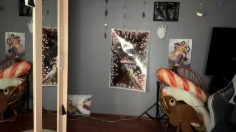 Media: Video of a cozy bedroom with two wicker chairs adorned with plush toys and blankets. Gray walls feature framed posters and string lights, creating a whimsical, childlike atmosphere.