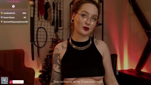 Media: Video of a fair-skinned, blonde woman with tattoos, wearing glasses, a black crop top, and a silver choker, standing in a dimly lit room with BDSM gear and Christmas decorations.