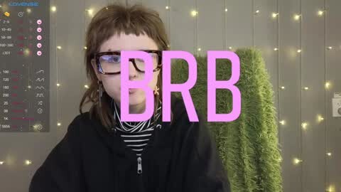 Media: Video of a young person with shoulder-length brown hair, wearing black glasses and a black hoodie, standing in front of a wall covered in green string lights. Text overlay reads \"BBB.\