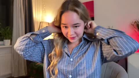 Media: Video of an Asian woman with long, wavy blonde hair, wearing a blue striped shirt, adjusting her hair in a living room with a lamp, potted plants, and a red wall.