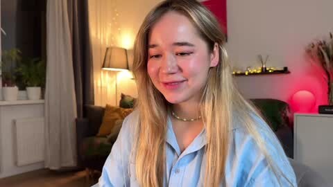 Media: Video of a young, light-skinned woman with long blonde hair, wearing a light blue button-up shirt and pearl necklace, smiling in a warmly lit, cozy living room with a green couch, potted plants, and modern decor.