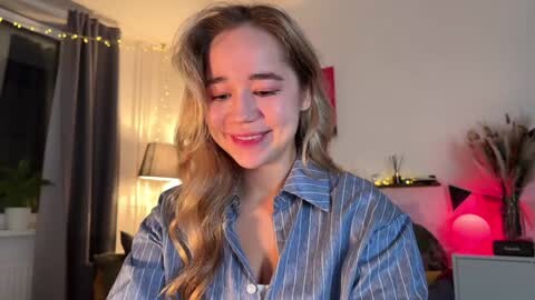 Media: Video of a smiling, light-skinned woman with long, wavy blonde hair, wearing a blue pinstriped button-down shirt. Background features a lit lamp, potted plants, and a vase of dried flowers.