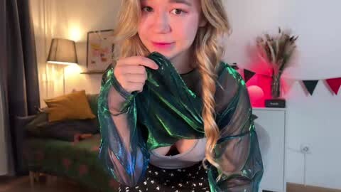 Media: Video of a young woman with wavy blonde hair, wearing a sheer green blouse, white bra, and polka-dot skirt, standing in a cozy living room with a green couch, lamp, and festive decorations.