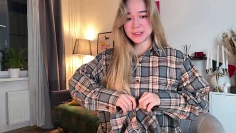 Media: Video of a young woman with long blonde hair, smiling, wearing a plaid shirt, sitting on a couch in a cozy, warmly lit living room with a green cushion, lamp, and potted plants.