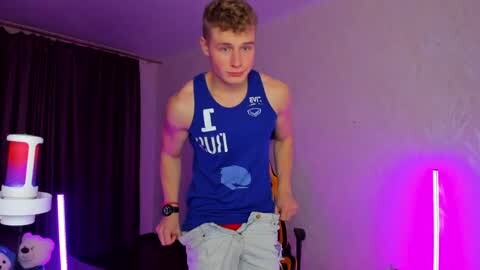 Media: Video of a young, fit man with short, light brown hair, wearing a blue tank top and light jeans, in a dimly lit room with purple lighting, holding a red cup.