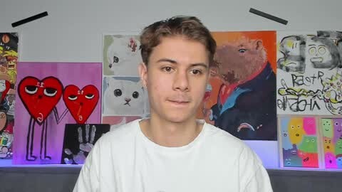 Media: Video of a young man with short brown hair in a white shirt, standing in front of a wall adorned with colorful, abstract art and graffiti.