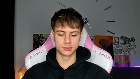Media: Video of a young Caucasian male with short, brown hair, wearing a black hoodie, seated in a pink and white gaming chair with \"SOMEONE\" text. Background features a cluttered room with colorful posters and a clock.