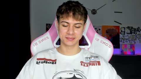 Media: Video of a young man with short brown hair wearing a white shirt with logos, seated in a pink and white gaming chair. Background features a colorful, abstract wall painting and a black door.