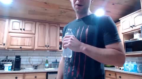 Media: Video of a muscular man in a dark t-shirt, holding a water bottle in a modern kitchen with light wood cabinets and white subway tiles.