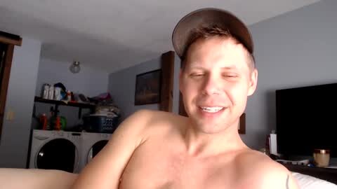 Media: Video of a smiling, shirtless, young white man with short brown hair and a baseball cap, standing in a laundry room with shelves and a flat-screen TV.
