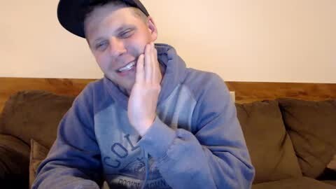 Media: Video of a smiling, fair-skinned man with short hair, wearing a black cap and gray hoodie, resting his hand on his cheek while sitting on a brown couch against a plain white wall.