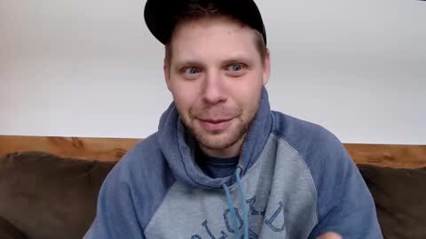 Media: Video of a smiling white man with short hair, wearing a blue baseball cap and hoodie with a grey logo. He sits on a brown couch against a white wall.