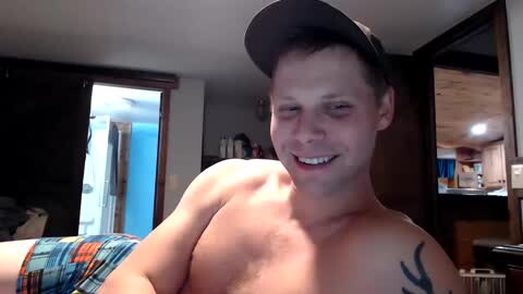 Media: Video of a shirtless, smiling young man with a cap and a tattoo, standing in a dimly lit, cozy room with wooden furniture and a blue door.