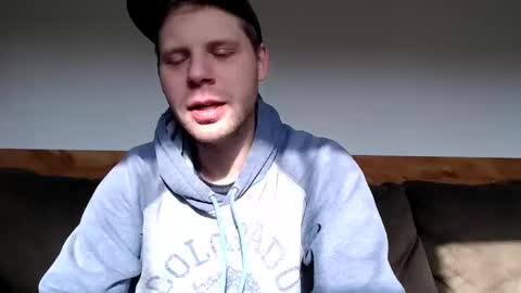 Media: Video of a young Caucasian man with short brown hair, wearing a black baseball cap and blue hoodie, sitting on a brown couch, eyes closed, possibly sleeping.