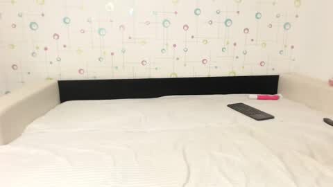 Media: Video of a minimalist bedroom featuring a white bed with a black headboard, white bedding, a pink remote, and a white wall adorned with colorful, abstract circular patterns.