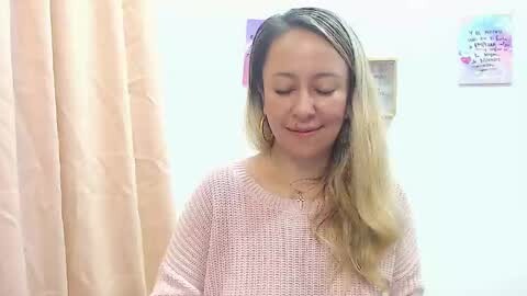 Media: Video of a young woman with long, wavy blonde hair, wearing a light pink knit sweater, standing in a room with beige curtains and a motivational poster on the wall.
