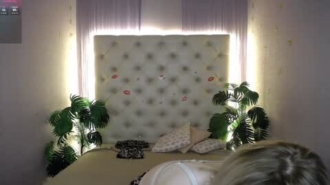 Media: Video of a cozy bedroom with a tufted headboard, lit from below, featuring green leafy plants, pillows, and a woman's blonde hair, with a soft, romantic ambiance.