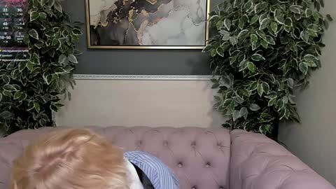 Media: Video of a light-haired child sitting on a plush, light pink sofa, with a framed map hanging on a dark gray wall. Two large, green potted plants flank the sofa.