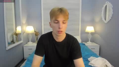 Media: Video of a young man with short blonde hair, wearing a black t-shirt, sitting on a blue bed in a small, dimly-lit room with white walls, two nightstands, and a mirror.