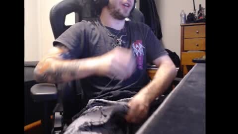 Media: Video of a man with a beard, wearing a headset, gray shirt, and shorts, sitting in a gaming chair, playing a video game.