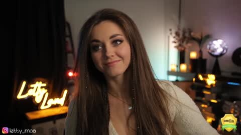 Media: Video of a young woman with long brown hair and fair skin, wearing a white lace top, smiling softly, in a dimly-lit, cozy room with candles, a \"Let It Snow\" sign, and a social media watermark.
