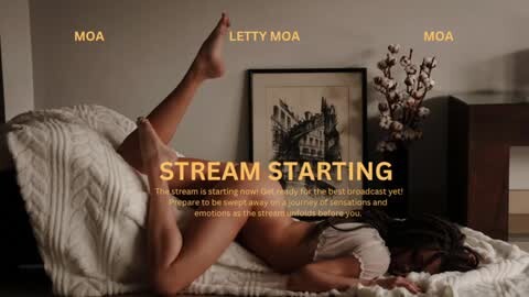 Media: Video of a nude woman with fair skin and dark hair, lying on her stomach on a textured white blanket, legs raised, in a dimly lit room with a framed black-and-white photo of a cityscape on the wall.