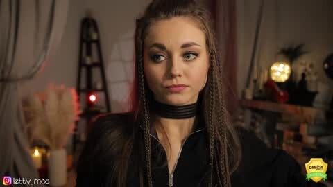 Media: Video of a young woman with long, braided hair and a black choker, standing indoors, surrounded by dimly lit decor, including a ladder and a table with candles.