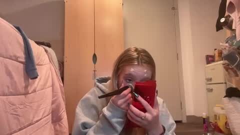 Media: A candid video of a young girl with fair skin and blonde hair, wearing a light grey robe, applying makeup in a cluttered bathroom.
