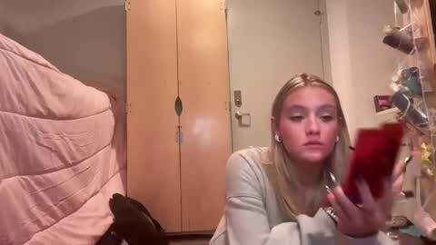 Media: Video of a young blonde woman with fair skin, wearing a light sweater, looking at a red phone, in a messy bedroom with wooden doors and a pink quilted bedspread.