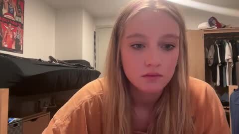 Media: Video of a young blonde girl with fair skin, wearing an orange shirt, sitting on a bed in a cluttered bedroom with a black bedspread, a wooden wardrobe, and a red London-themed poster on the wall.