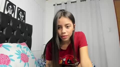 Media: Video of a young Latina woman with long black hair, wearing a red t-shirt, sitting on a bed with pink and blue floral sheets.