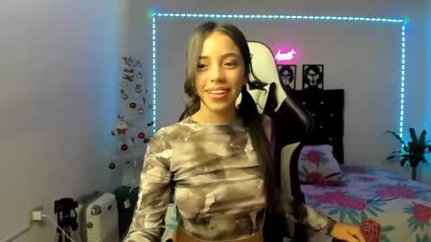 Media: Video of a smiling woman with medium-length, dark hair, wearing a camo-patterned crop top, seated in a gaming chair, with a neon-lit room in the background.