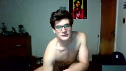 Media: Video of a shirtless young man with glasses, medium build, short brown hair, sitting on a bed in a dimly lit room. Background includes a dresser, a door, and a colorful poster.