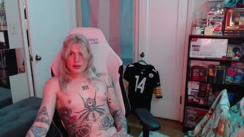 Media: Video of a topless, tattooed, blonde woman with small breasts, sitting in a gaming chair, in a room filled with gaming memorabilia and toys.