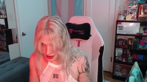 Media: Video of a young woman with platinum blonde hair, wearing a white lace top, sitting in a black and white gaming chair. She has tattoos on her arm and shoulder. The background features a colorful game shelf and a pink and blue curtain.