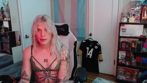 Media: Video of a young, blonde woman with tattoos, wearing a green lace bralette, seated on a gaming chair in a cluttered, colorful room with sports jerseys and toys.
