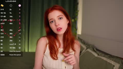 Media: A video of a pale-skinned, red-haired woman in a beige dress, sitting on a bed with green curtains in the background.