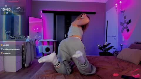 Media: Video of a young woman with fair skin and long blonde hair, wearing a gray sports bra and sweatpants, kneeling on a bed in a dimly lit bedroom with neon pink and blue lights.