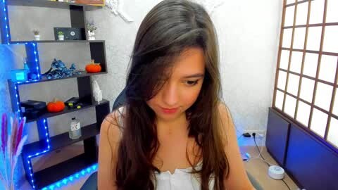 Media: A video of a young woman with long dark hair, wearing a white top, sitting in a dimly lit room with white walls, a black bookshelf, and blue LED strip lights.
