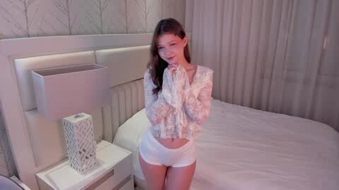 Media: A video of a young woman with long brown hair, wearing a lace top and white shorts, standing in a modern bedroom with white walls and a white bed.