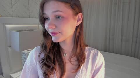 Media: Video of a young Caucasian woman with long, wavy brown hair, wearing a white blouse, in a softly lit bedroom with white walls and a white bed.
