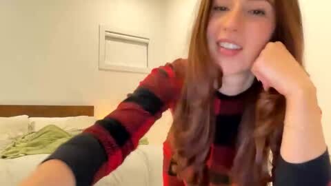 Media: Video of a young woman with long brown hair, smiling, wearing a red and black striped shirt, sitting on a bed with white sheets and green pillows in a simple, brightly lit room.