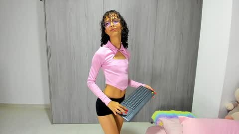 Media: Video of a slender, young woman with curly black hair and glasses, wearing a pink long-sleeve crop top and black shorts, holding a laptop, standing in a bedroom with light-colored walls and a gray wardrobe.
